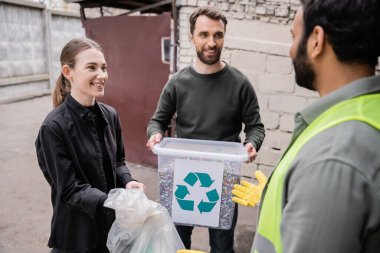 Açık hava atık imha istasyonunda, çöp ayıklama ve geri dönüşüm konseptinde, güvenlikli yelek ve eldiven giyen bulanık Hintli işçilerin yanında gülen gönüllüler