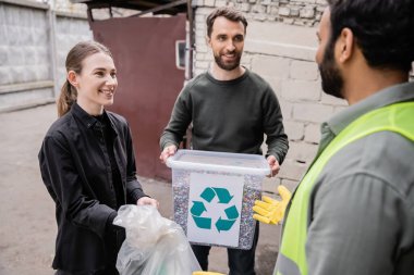 Smiling volunteers holding waste near blurred indian worker in safety vest and glove in outdoor waste disposal station, garbage sorting and recycling concept clipart