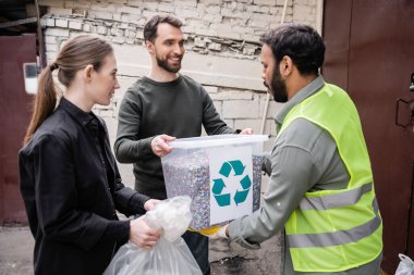 Smiling volunteers giving trash to indian worker in high visibility vest in outdoor waste disposal station, garbage sorting and recycling concept clipart