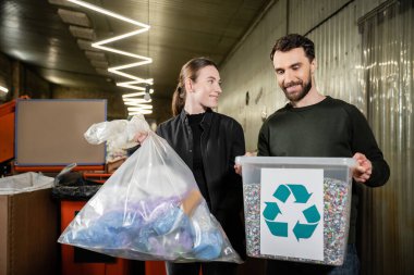 Smiling volunteer holding trash bag near man with bin and recycle sign in blurred waste disposal station at background, garbage sorting and recycling concept clipart