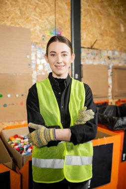Yüksek görüş mesafesi olan yelek ve eldivenlerle gülümseyen işçiler kameraya bakıyor ve kollarını çaprazlarken aynı zamanda çöp öğütme istasyonunda bulanık plastik kapsüllerin yanında duruyor, çöp ayıklama ve geri dönüşüm konsepti
