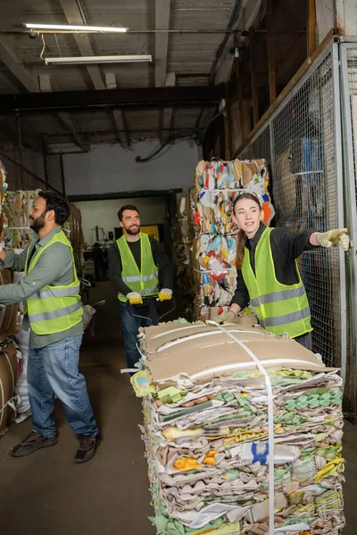 stock image Smiling female worker in reflective vest and gloves pointing away while standing near waste paper and colleagues with hand pallet truck in garbage sorting center, waste sorting and recycling concept