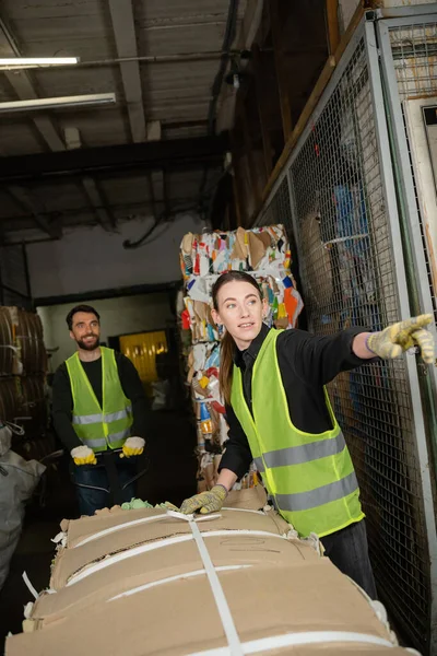 worker in protective vest and gloves pointing with finger away while standing near waste paper and colleague with hand palette truck in garbage sorting center, waste sorting and recycling concept