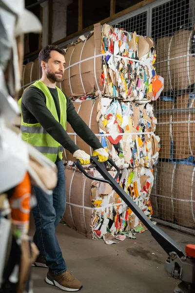 Bearded male worker in protective vest and gloves using hand pallet truck while standing near waste paper in garbage sorting center, waste sorting and recycling concept