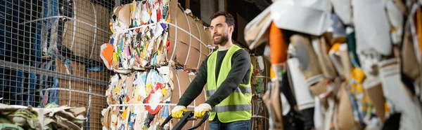 Bearded sorter in high visibility vest and gloves standing near hand pallet truck while standing near waste paper in blurred garbage sorting center, waste sorting and recycling concept, banner