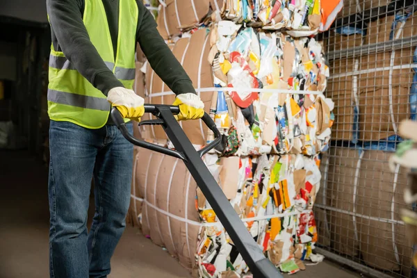 Cropped view of male sorter in protective gloves and vest using hand pallet truck while working near waste paper in blurred garbage sorting center, waste sorting and recycling concept