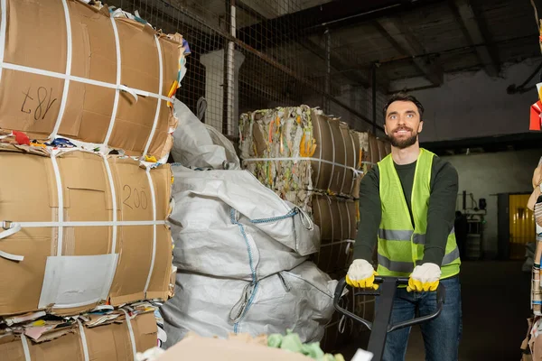 Smiling and bearded worker in reflective vest and gloves using hand pallet truck while moving waste paper in blurred garbage sorting center, waste sorting and recycling concept