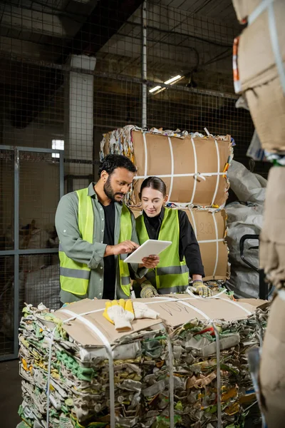 stock image Indian worker in safety vest using digital tablet near female colleagues while standing near waste paper on hand pallet truck in waste disposal station, garbage sorting and recycling concept