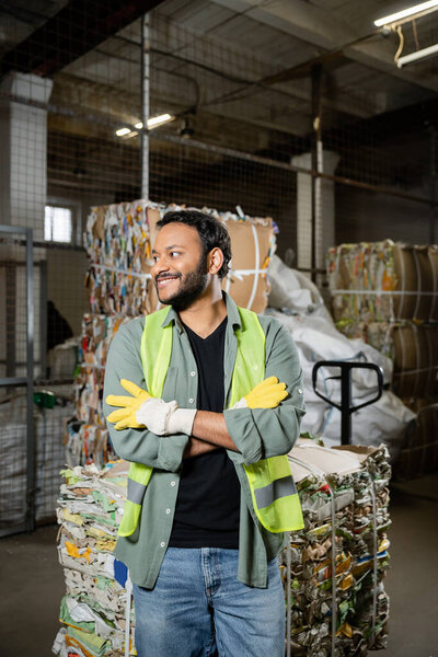 Positive indian sorter in protective gloves and vest crossing arms while standing near waste paper on hand pallet truck in blurred waste disposal station, garbage sorting and recycling concept