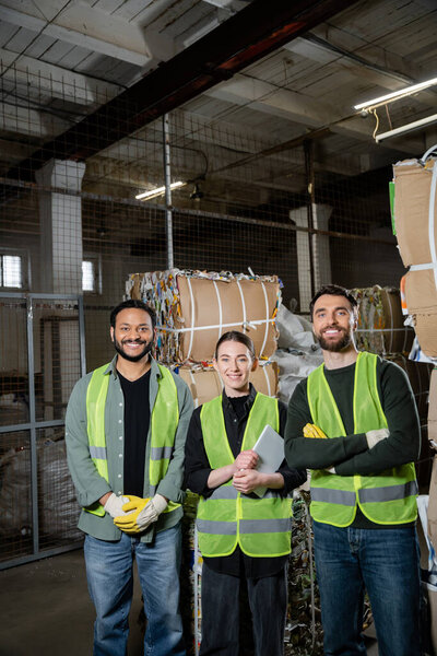Smiling interracial sorters in protective vests holding digital tablet and looking at camera near waste paper in waste disposal station, garbage sorting and recycling concept