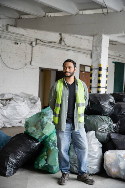 Bearded indian sorter in high visibility vest looking away while standing near plastic bags and blurred sacks while working in garbage sorting center, recycling concept