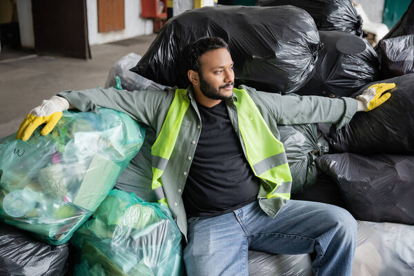 Bearded indian sorter in high visibility vest and gloves looking away while resting on plastic bags with trash in garbage sorting center, recycling concept