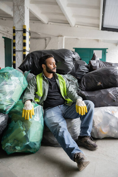 Indian worker in high visibility vest and gloves looking away while sitting and resting on plastic bags with trash while working in  garbage sorting center, recycling concept