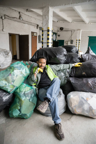 Confident indian worker in high visibility vest and gloves looking at camera while sitting on plastic bags with trash in garbage sorting center, recycling concept