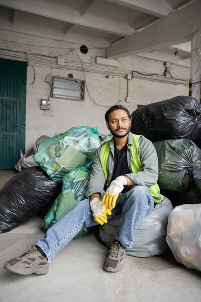 Male indian worker in high visibility vest and protective gloves sitting on plastic bags with trash while working in garbage sorting center, recycling concept
