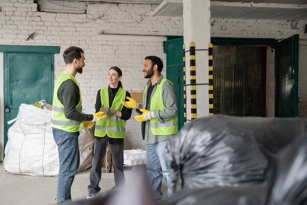 Smiling interracial workers in protective vests and gloves talking while standing and resting near blurred plastic bags with trash in garbage sorting center, recycling concept