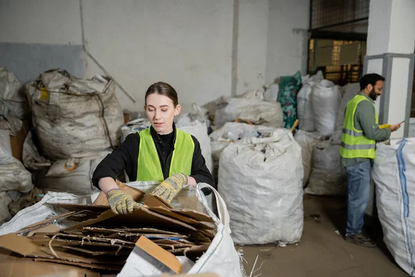 Young female worker in protective vest and gloves putting waste paper in sack while working near blurred indian colleague in waste disposal station, garbage sorting and recycling concept