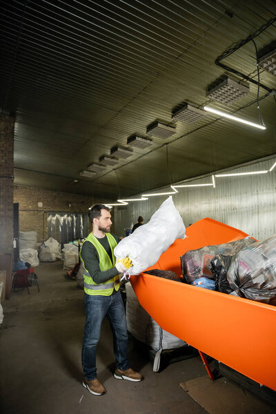 Side view of bearded male worker in glove and fluorescent vest putting sack with trash in container in blurred waste disposal station, garbage sorting and recycling concept