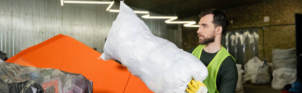 Side view of bearded man in protective vest and glove putting sack with trash in container while working in waste disposal station, garbage sorting and recycling concept, banner 