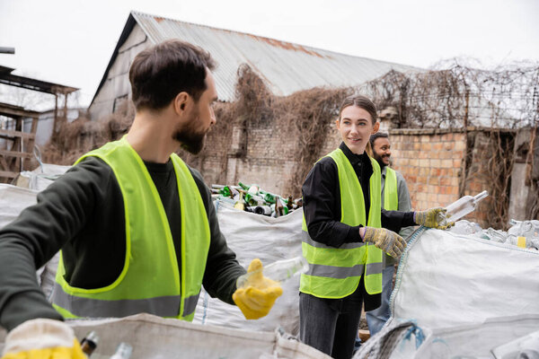 Smiling worker in safety vest and gloves holding glass trash near multiethnic colleagues and sacks in outdoor waste disposal station, garbage sorting and recycling concept