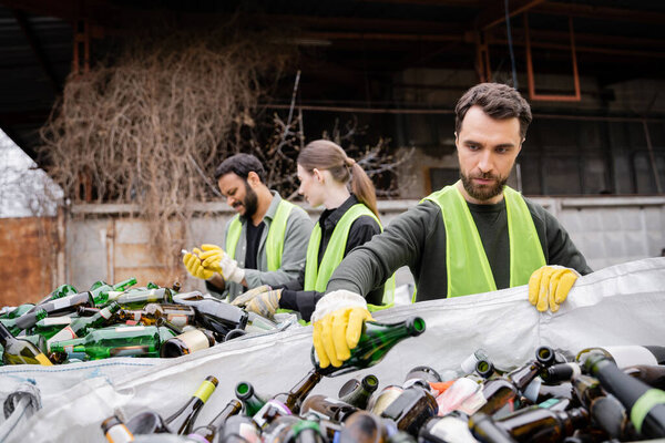 Bearded worker in protective gloves and vest putting glass bottle in sack near blurred interracial colleagues in outdoor waste disposal station, garbage sorting and recycling concept