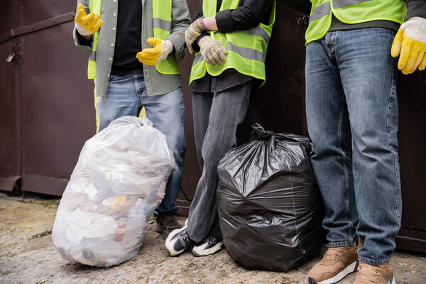 Cropped view of workers in high visibility vests and gloves standing near trash bags outdoors in waste disposal station, garbage sorting and recycling concept
