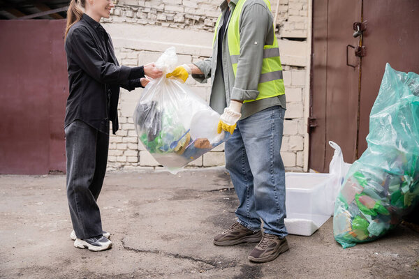 Cropped view of smiling volunteer giving plastic bag with trash to worker in protective vest and gloves near trash outdoors in waste disposal station, garbage sorting and recycling concept