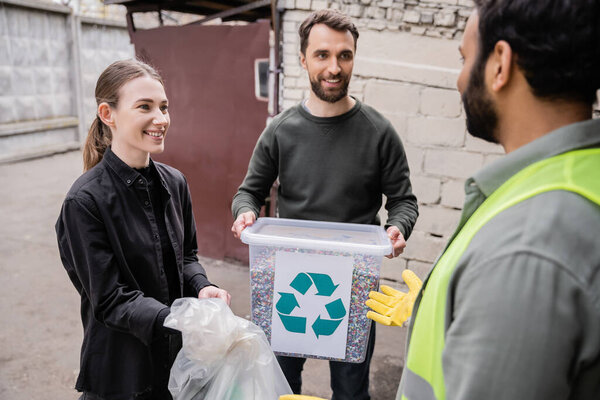 Smiling volunteers holding waste near blurred indian worker in safety vest and glove in outdoor waste disposal station, garbage sorting and recycling concept