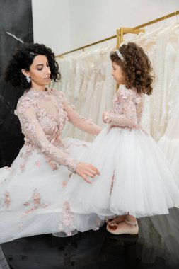 charming middle eastern bride with brunette wavy hair in wedding dress adjusting tulle skirt of daughter in cute floral attire in bridal salon, shopping, special moment, togetherness  clipart