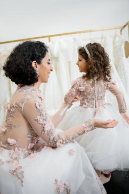 Kahverengi dalgalı saçlı, çiçekli gelinlikli mutlu ortadoğulu kadın gelinlik salonundaki tatlı elbiseli, alışveriş yapan, özel bir an, birliktelik içinde gülümseyen kızın tüllü eteğine bakıyor. 
