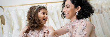 happy middle eastern woman in floral wedding dress hugging shoulders of cheerful girl in cute attire in bridal salon, shopping, special moment, mother and daughter, bonding, banner  clipart