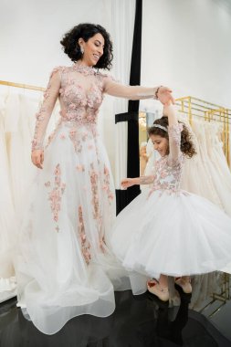 happy middle eastern woman in floral wedding dress dancing with smiling girl in cute attire with tulle skirt in bridal salon, shopping, special moment, mother and daughter, happiness  clipart