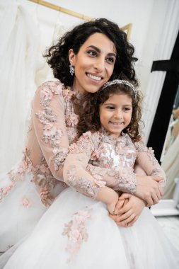 cheerful middle eastern woman in floral wedding dress hugging happy girl in cute attire in bridal salon, shopping, special moment, mother and daughter, happiness  clipart