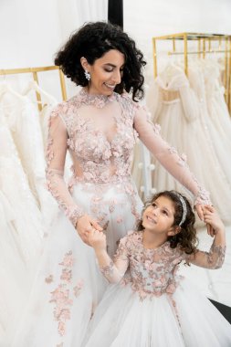 delightful middle eastern bride in floral wedding dress holding hands with happy girl in cute attire in bridal salon, shopping, special moment, mother and daughter, happiness, togetherness  clipart