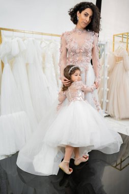 cute girl in floral attire hugging elegant woman with brunette hair standing in wedding dress near blurred white gown inside of luxurious bridal salon, shopping, bride-to-be, mother and daughter  clipart
