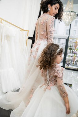 cute middle eastern girl in floral attire holding hands with woman and walking in floral dresses near blurred wedding gown inside of bridal salon, shopping, bride-to-be, mother and daughter  clipart