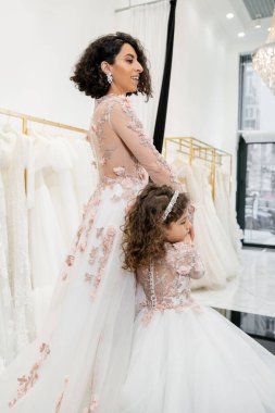 cute middle eastern kid in floral attire holding hands with happy woman and walking in floral dresses near blurred wedding gown inside of bridal salon, shopping, bride-to-be, mother and daughter  clipart