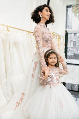 middle eastern girl in floral attire holding hands with cheerful mother standing in wedding dress near blurred white gown inside of luxurious bridal salon, shopping, bride-to-be, bonding  clipart