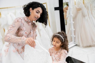 brunette middle eastern bride in floral wedding gown helping to choose dress for her cute little daughter in bridal boutique around white tulle fabrics, process of preparation, togetherness  clipart