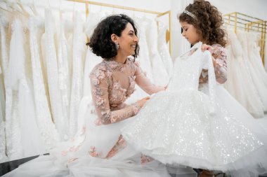 blissful and brunette middle eastern bride in floral wedding gown helping to choose dress for her cute little daughter in bridal salon around white tulle fabrics, process of preparation  clipart