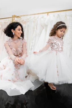 special moment, joyful middle eastern bride in floral wedding gown sitting and looking at little daughter in bridal salon posing around white tulle fabrics, bridal shopping, togetherness  clipart
