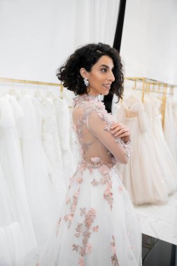 cheerful middle eastern and brunette woman with wavy hair standing in gorgeous and floral wedding dress inside of luxurious bridal salon around white tulle fabrics, bridal shopping, looking away  clipart