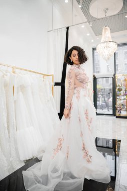 full length of middle eastern and brunette woman with wavy hair standing in gorgeous and floral wedding dress with train inside of luxurious bridal salon around white tulle fabrics, bridal shopping  clipart
