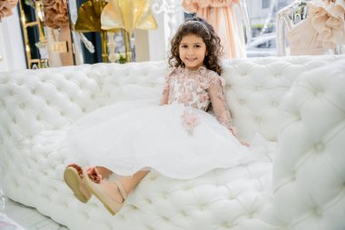 cheerful middle eastern girl with curly hair sitting in floral dress with tulle skirt and shoes on white couch inside of luxurious wedding salon, smiling kid, blurred background  clipart