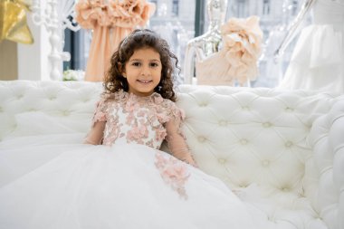 cheerful middle eastern girl with curly hair sitting in floral dress on white couch and smiling inside of luxurious wedding salon, smiling kid, tulle skirt, blurred background  clipart