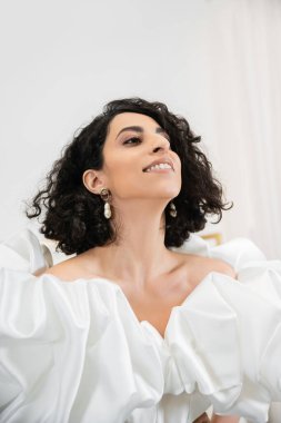Orta Doğulu mutlu gelinin portresi esmer ve dalgalı saçlı moda gelinlik içinde poz veren kabarık kollu ve fırfırlı gelinlik, beyaz gelinlik.
