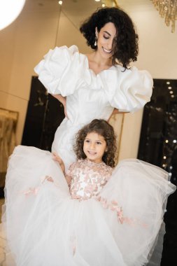 happy little girl in floral attire holding tulle skirt and looking away near  middle eastern mother with wavy hair standing in white wedding dress with puff sleeves and ruffles in bridal boutique  clipart