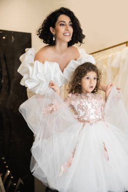 captivating middle eastern bride with brunette hair standing in white wedding gown with puff sleeves and ruffles and looking away while holding tulle skirt of surprised daughter in bridal store  clipart