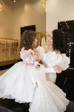 Güzel bir ortadoğulu gelin, esmer saçlı, beyaz gelinlik içinde, kabarık kolları ve fırfırları olan, gelinlik butiğinde tül etekli, kız gibi giyinen bir kızın yanında duruyor.  