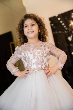 cheerful middle eastern flower girl in floral dress with tulle skirt standing with hands on hips and looking away in bridal boutique, preparation for wedding, blurred background  clipart